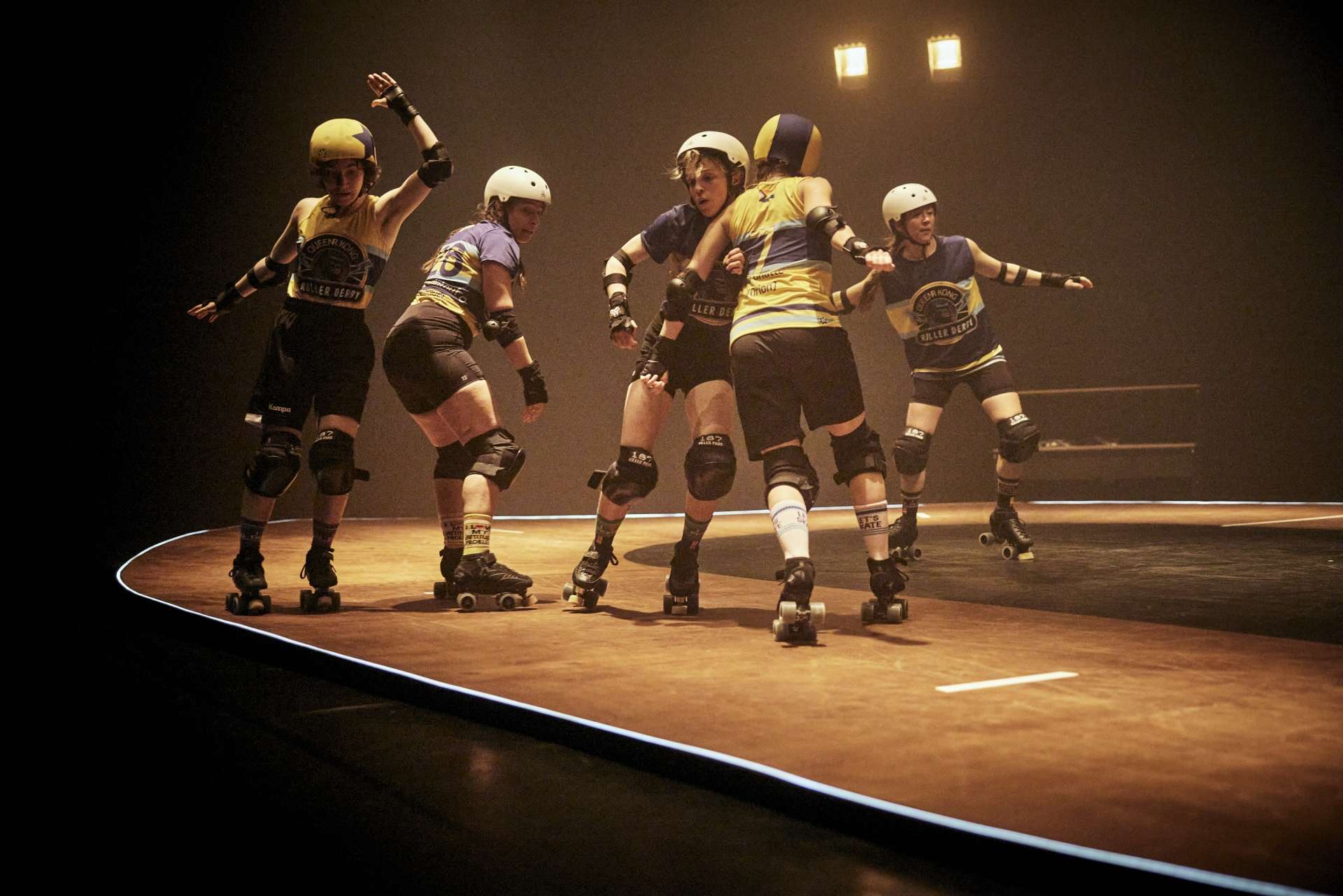 Sur un mini track en bois, cinq interprètes pratiquent le roller derby. Elles ont des maillots jaune et bleu.