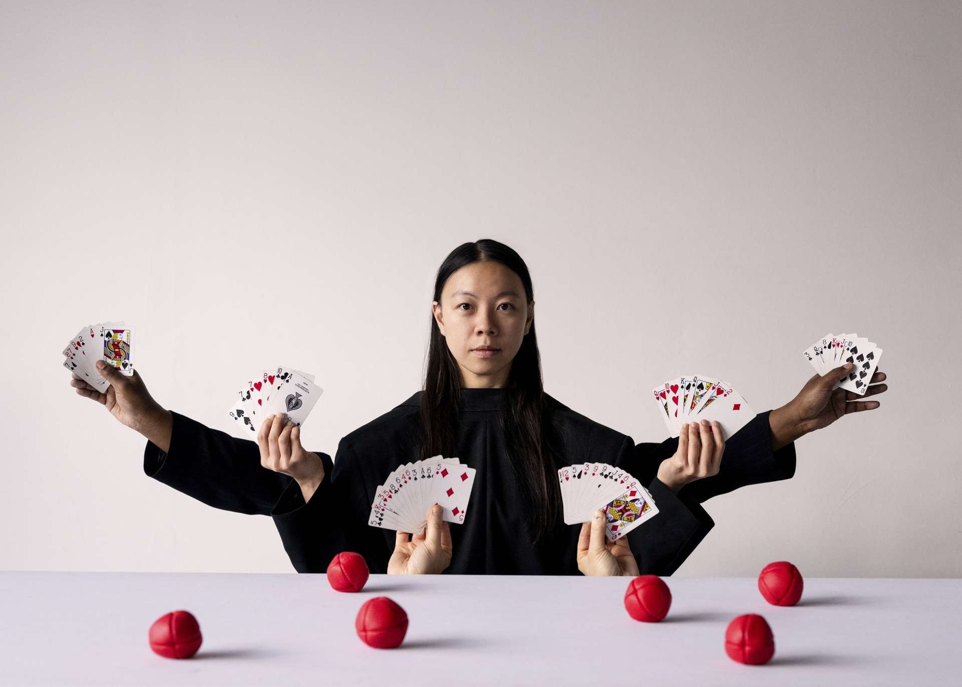 De face, une femme en haut de costume noir. Elle est assise et sur le fond blanc ressortent cartes de jeu et balles de jonglages rouges. Les éléments apparaissent autour d'elle en mode Shiva. 