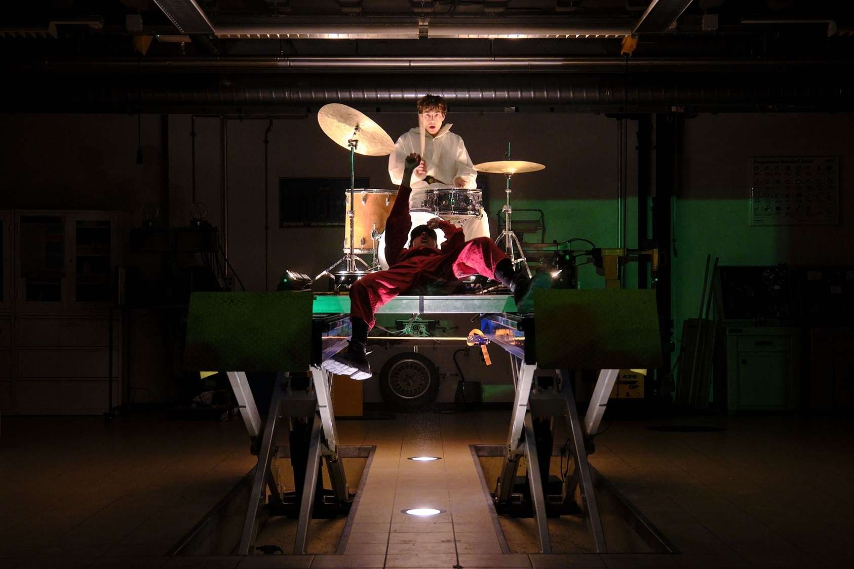 Dans une salle, un musicien joue de la batterie en hauteur sur une table. Une performeuse est allongée sur la table devant lui, son poing levé. 